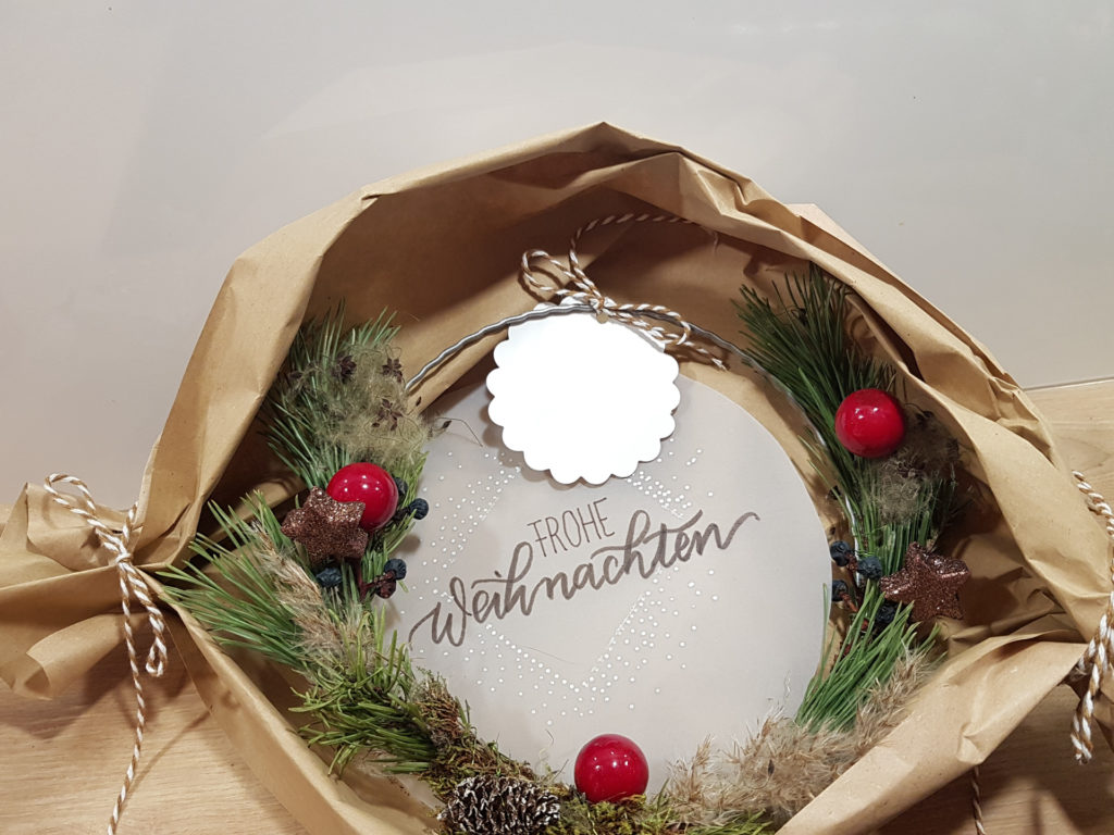 Kleiner selbstgebundener Weihnachtskranz mit lila Christbaumkugeln. Im Hintergrund steht auf Transparentpapier "Frohe Weihnachten".