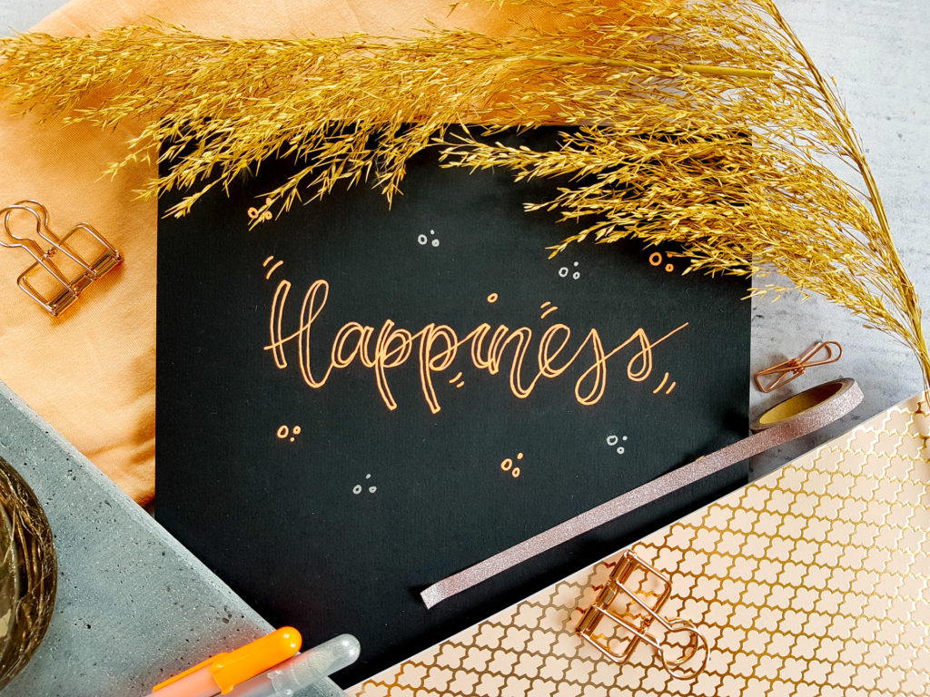 Ein Lettering mit dem Wort "Happiness" mit orange-leuchtenden Stiften im Faux-Calligraphie-Style auf schwarzem Papier