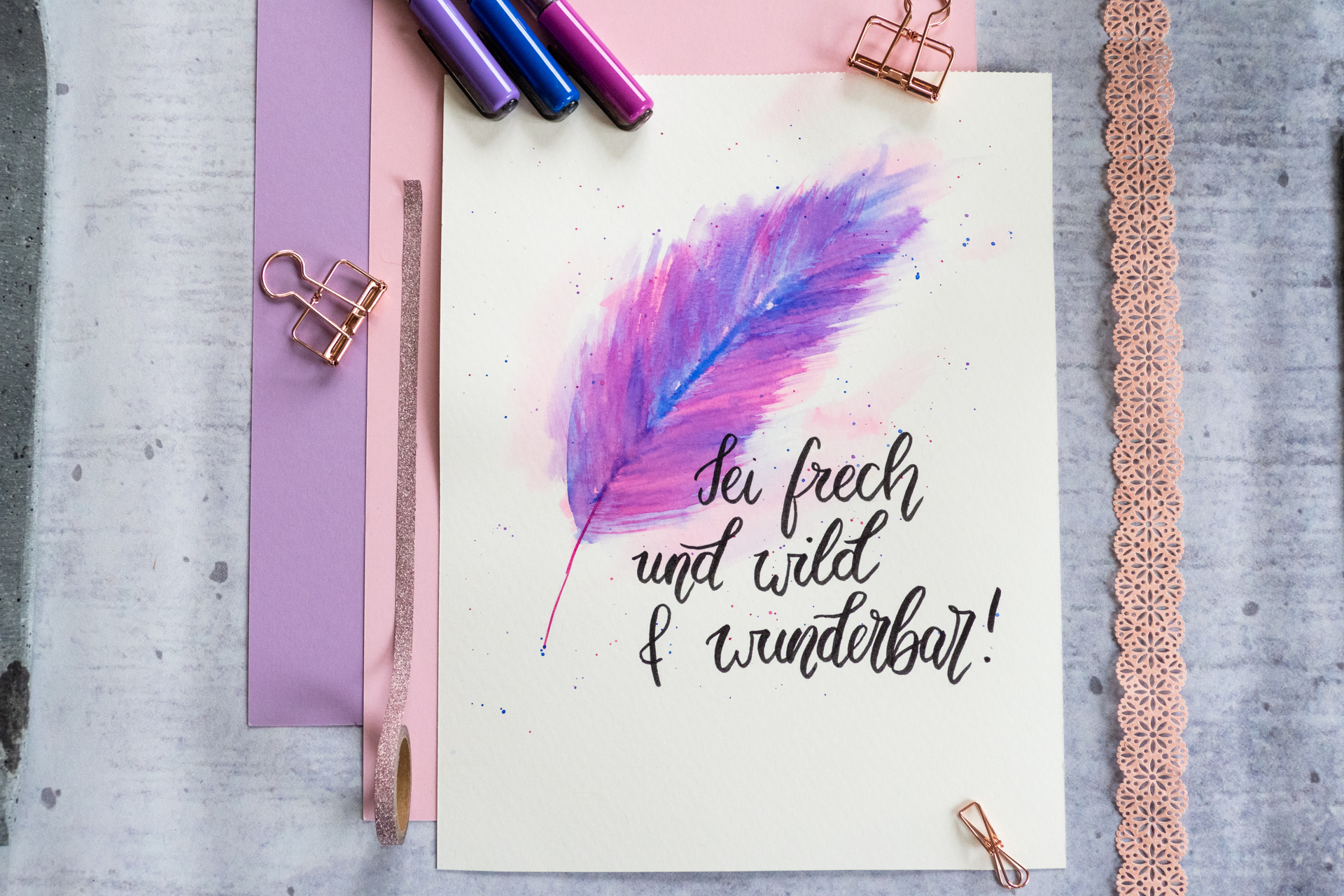 Spruch "Sei frech und wild und wunderbar" mit schwarzem Brushpen geschrieben. Im Hintergrund ist eine Feder im "Watercolor-Style" in den Farben blau und lila.
